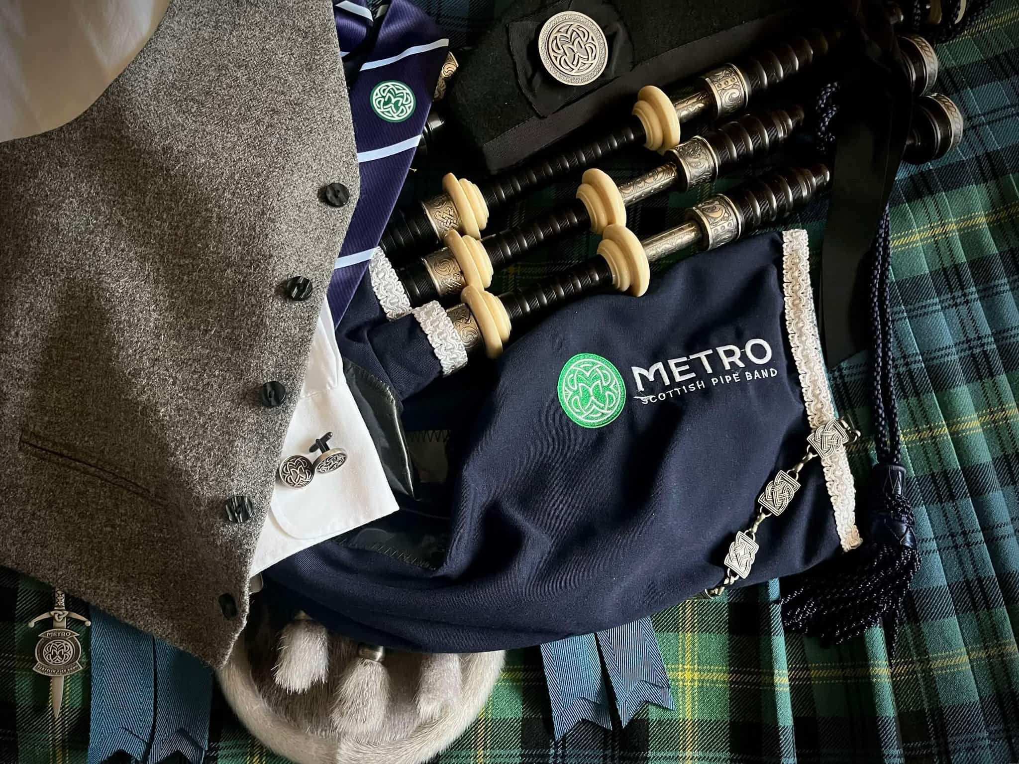 Metro Scottish Pipe Band logo embroidered on a navy bagpipe cover, laid on a tartan kilt with a grey waistcoat and silver kilt pin